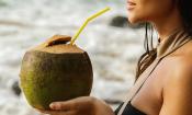 Junge Frau in Badekleid trinkt Kokoswasser mit Strohhalm direkt aus Kokosnuss.