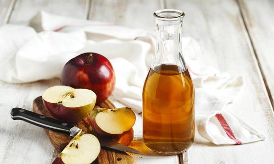 Apple cider vinegar in a glass carafe, on the right a wooden board with an apple and a sliced apple.