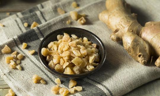 Jengibre confitado en un recipiente negro junto a algunos trozos de jengibre y al tubérculo entero.