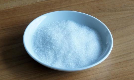 Granulated sugar heaped in a white plate on wooden base.