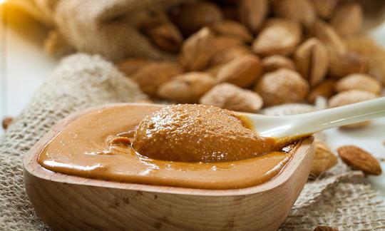 Almond butter, pure, without additional salt in a rounded wooden bowl, behind it sack of almonds.