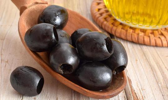Черные оливки, нарезанные деревянными ложками, рядом со стеклянной бутылкой с оливковым маслом.