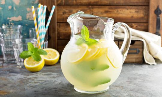 Lemon juice, raw: The glass pitcher with a handle contains lemon juice diluted with water. In the background to the left are two lemon halves and  glasses.