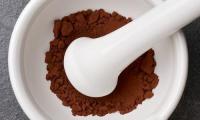 Kakaopulver, roh, ungesüsst in einem Mörser oder Reibschale aus weisser Keramik mit Stössel.