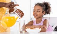 A woman pours orange juice into a girl's drinking glass.