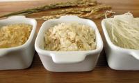 Three white bowls: rice (left), rice flakes (middle), rice noodles (right), and rice plants (back)