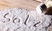Соль (столовая соль) высыпанная из солонки на деревянном столе. Мы употребляем слишком много соли..