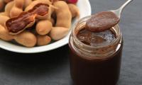 Tamarind paste in a glass container, to the left of it entire tamarinds, one of them opened.