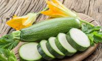 Zucchini—Curcubita pepo: Zucchini on a wooden board with slices of the vegetable in the foreground and zucchini blossoms in the background.